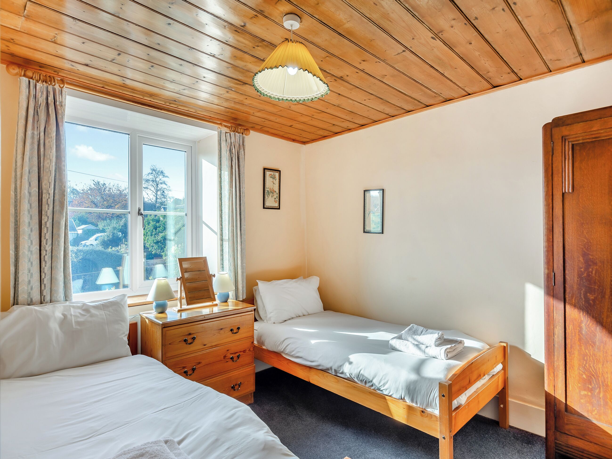 A bedroom with two beds, a wooden dresser, a window with curtains, and a wooden ceiling.