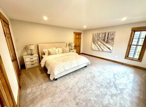 A bedroom with a large bed, a nightstand, a lamp, a wooden door, and a window.