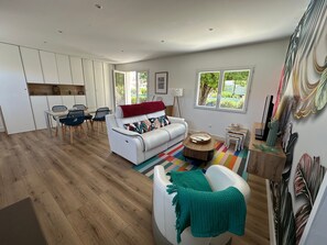 A modern living room with a white sofa, a dining area with chairs, and a kitchen area with cabinets. - Sous le Marronnier - Maison pour 4 avec jardin (Royan)