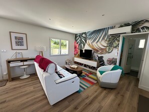 A modern living room with a white sofa, a TV, a desk, and a large tropical leaf mural. - Sous le Marronnier - Maison pour 4 avec jardin (Royan)