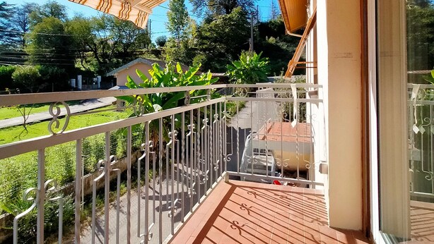 A balcony with a tiled floor and metal railing, overlooking a garden and a street.
