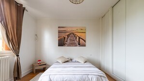 A bedroom with a wooden floor, a bed with white and gray striped bedding, a small bedside table with a red object, and a painting of a pier on the wall.
