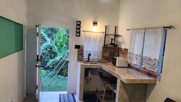 Habitación pequeña con cama, escritorio y silla. Tiene ventana con cortinas, lámpara colgante y vistas a la vegetación exterior.