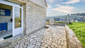 A stone house with a kitchen visible through a glass door, a stone patio, and a view of a village in the distance. - La Tour Vauban - Vue sur le port (Port-en-Bessin-Huppain)