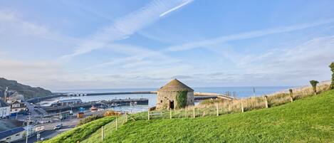 Een kustlandschap met een grasheuvel, een stenen gebouw en een haven met boten.