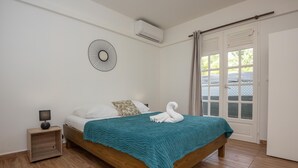 A bedroom with a wooden bed, a teal blanket, a white towel, a nightstand with a lamp, and a wall-mounted air conditioner. - L'Essentiel du KM22 - Appt avec terrasse et jardin (Petit-Bourg)