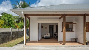 A house with a covered porch, a dining area, and a bedroom visible through an open door. - L'Essentiel du KM22 - Appt avec terrasse et jardin (Petit-Bourg)