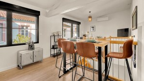 A dining area with a wooden table, four chairs, a small cabinet, and a window with a view of buildings. - Costa Del Sol - Charmant appt pour 2 avec terrasse (Toulouse)
