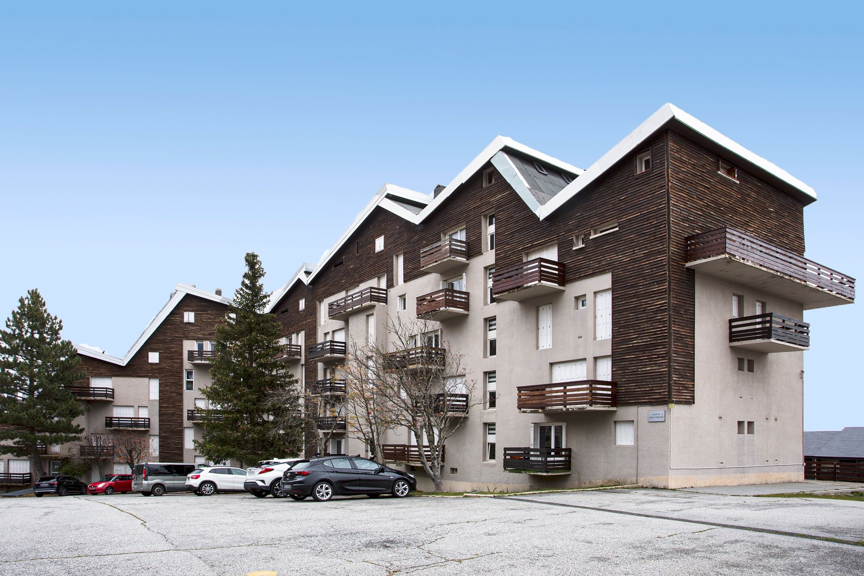 A multi-story residential building with balconies, a parking area, and a clear sky.