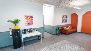 Un espacio de oficina moderno con un escritorio blanco, una máquina de café, una alfombra colorida, un sofá con cojines y una planta en maceta.