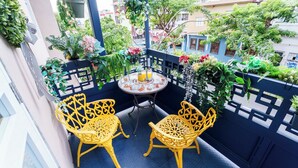 Een balkon met twee gele stoelen, een klein rond tafeltje met een drankje, en verschillende plantenpotten.