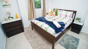 Een net opgemaakt bed met een kleurrijk dekbed, een nachtkastje met een lamp en een kleine potplant.