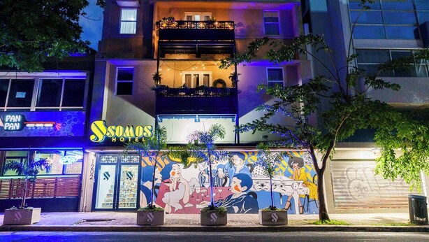 Een hotel met een muurschildering op de gevel, een bord met 'SOMOS HOTELS' en een restaurant genaamd 'PAN PAN' aan de overkant van de straat.