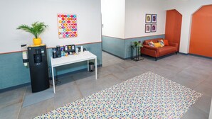 Ein moderner Arbeitsbereich mit einem weißen Tisch, einer Kaffeemaschine, farbenfroher Wandgestaltung, einer Couch und einem gemusterten Teppich.