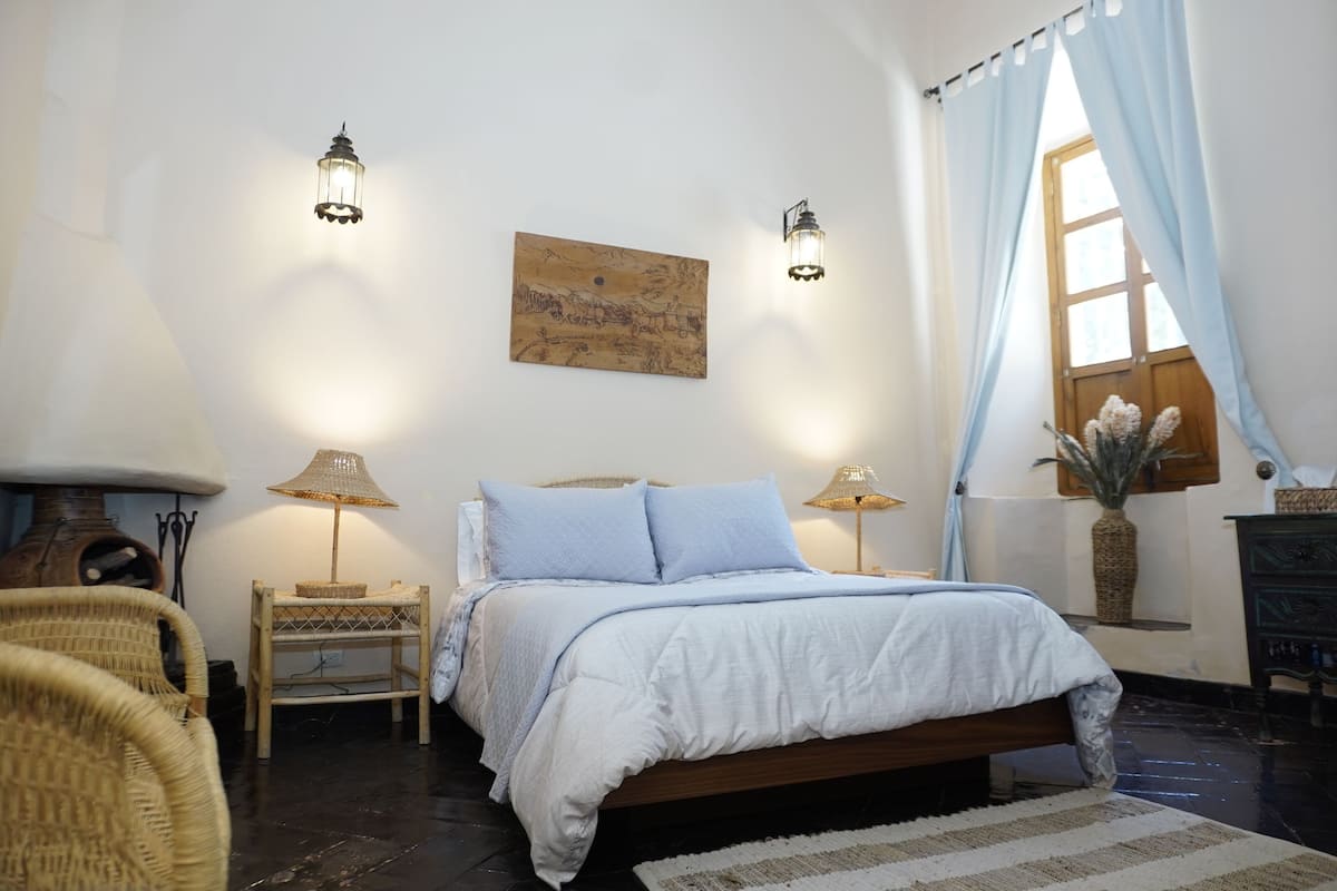 Een slaapkamer met een bed, twee bedlampen, een nachtkastje, een rieten stoel, een schilderij aan de muur en een raam met gordijnen.