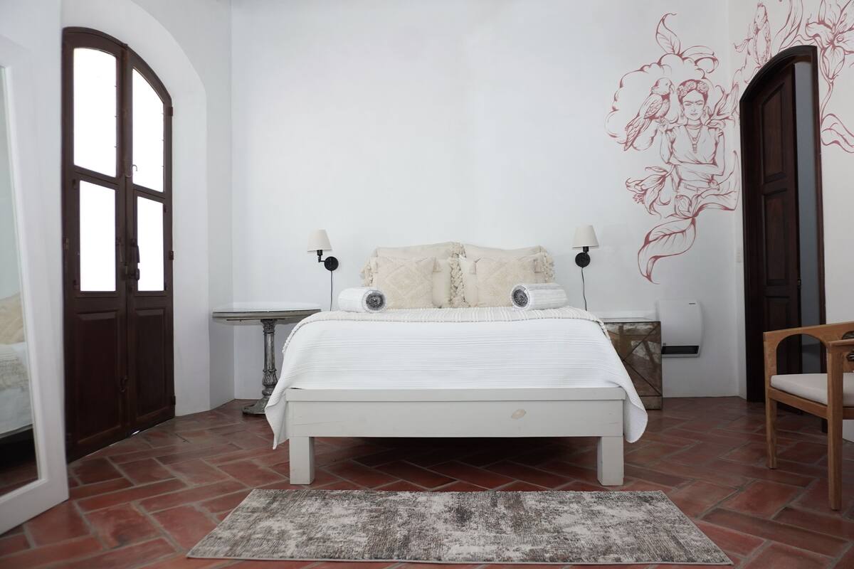 Een slaapkamer met een wit bed, een houten stoel, een klein tafeltje en een muur met een decoratief muurschildering.