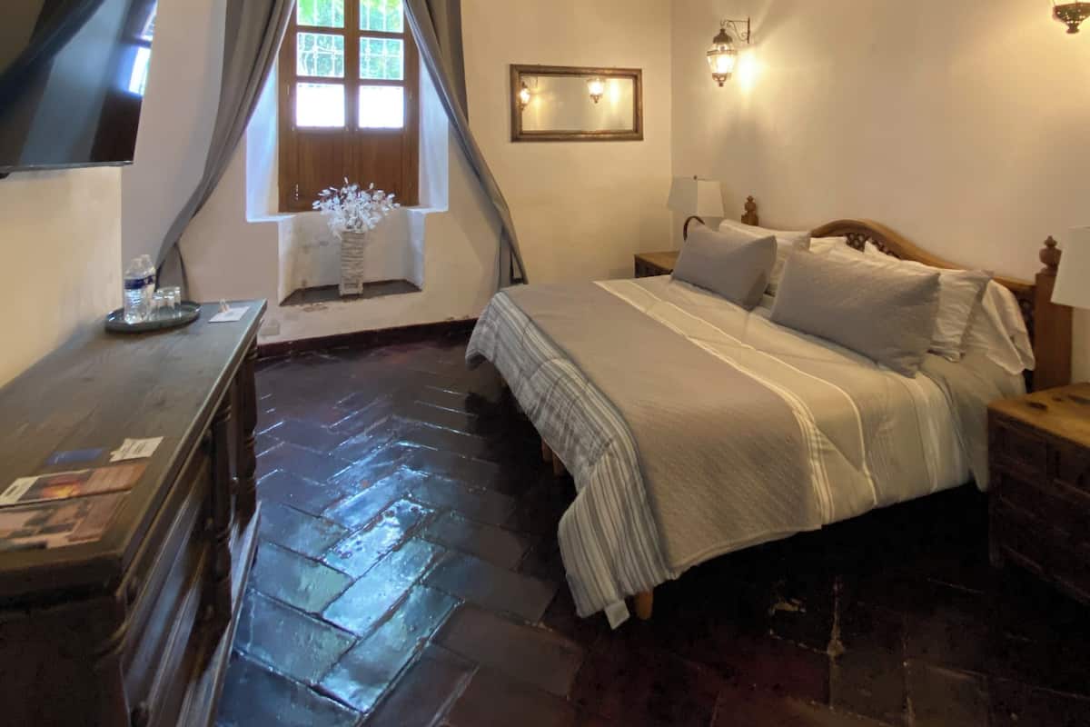 Een slaapkamer met een bakstenen vloer, houten balken, een kroonluchter, een bed met kussens, een nachtkastje, een aan de muur bevestigde televisie en een raam met gordijnen.