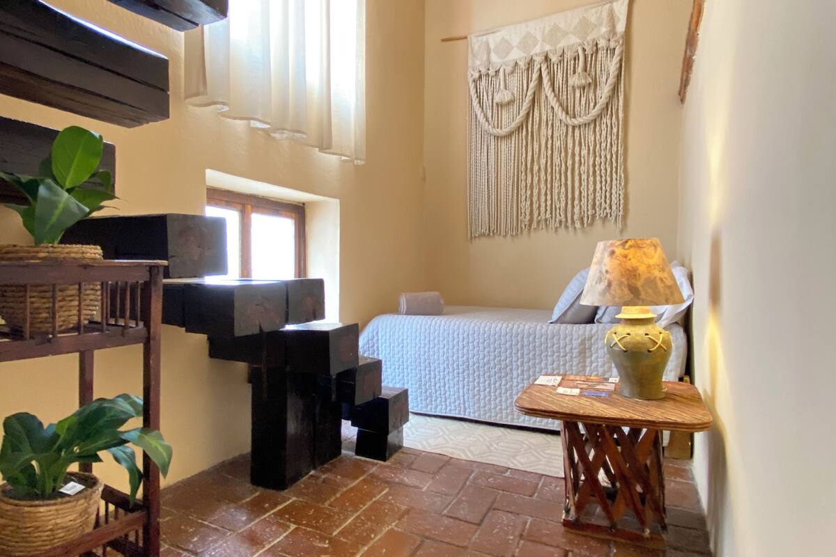 Een kleine, knusse kamer met een bed, een macramé wanddecoratie, een lamp en een plant op een plank.
