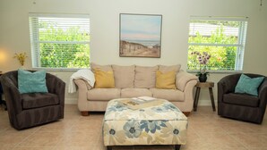 Una sala de estar con un sofá color crema, dos sillones, un taburete estampado y un cuadro en la pared.