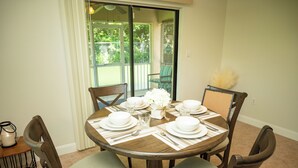 Mesa de comedor dispuesta para cuatro personas, con sillas, un jarrón de flores y vista al exterior a través de una puerta corrediza de vidrio.