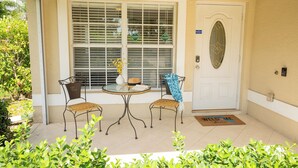 Un patio cubierto con una mesa redonda y dos sillas, una puerta con felpudo, y una ventana con persianas.