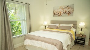 Un dormitorio ordenado con una cama, dos mesitas de noche con lámparas, una ventana con cortinas y una obra de arte floral en la pared.