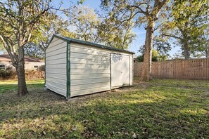 A small, single-story shed with a gabled roof, situated in a grassy yard with trees and a wooden fence in the background. - Charming 3-Bedroom Short-Term Rental in Port Neches (port neches)