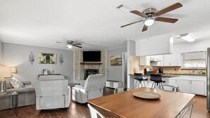 Een moderne woonkamer met een open haard, grijze fauteuils, een houten eettafel en een keuken met witte kasten en een zwart aanrecht.