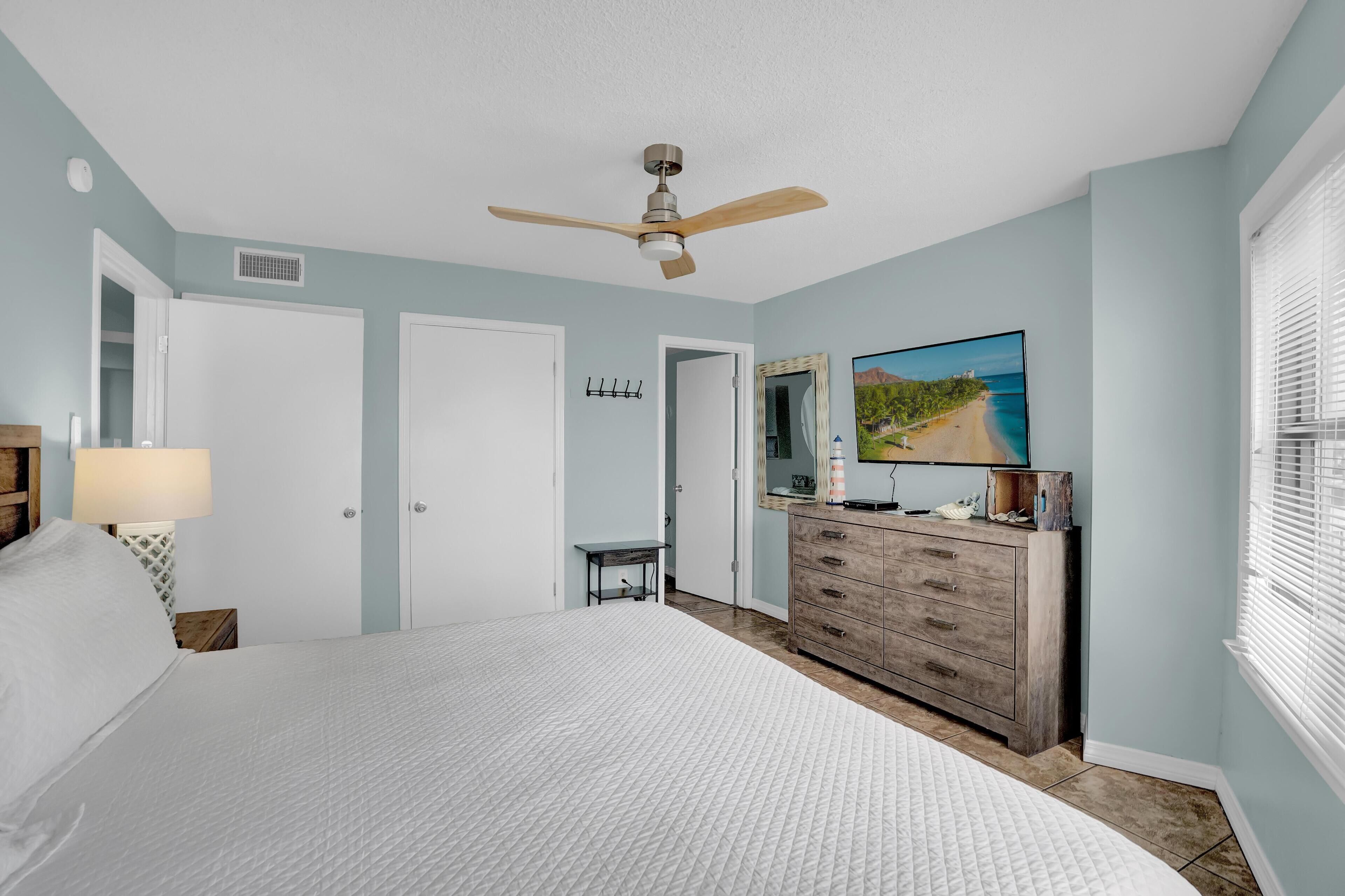 A bedroom with a bed, a dresser, a ceiling fan, a TV, and a window with blinds.