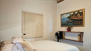 A bedroom with a bed, a window with blinds, a wall-mounted fan, a shelf with a plant, and a painting of a coastal scene. - Charming 2-bedroom house with WiFi, AC in Dibulla, ocean view. (Dibulla)