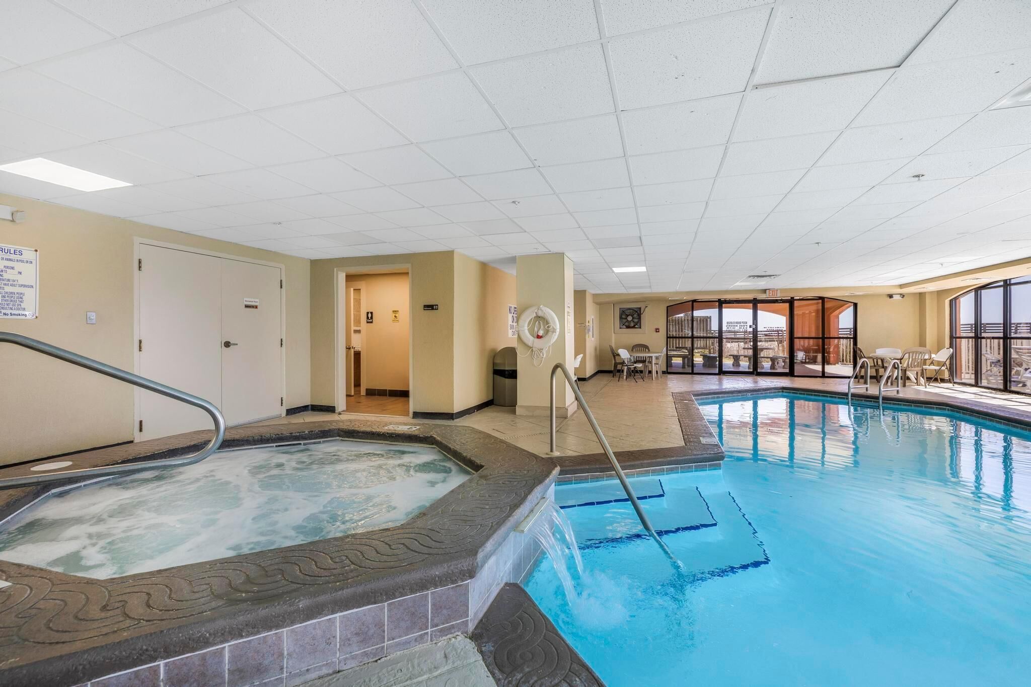 En indendørs swimmingpool med jacuzzi, et solbelyst område med stole og udsigt gennem store vinduer.