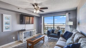A living room with a ceiling fan, a flat-screen TV, a sofa, a coffee table, and a large window offering a view of the ocean. - Harbour Place 503 (Orange Beach)