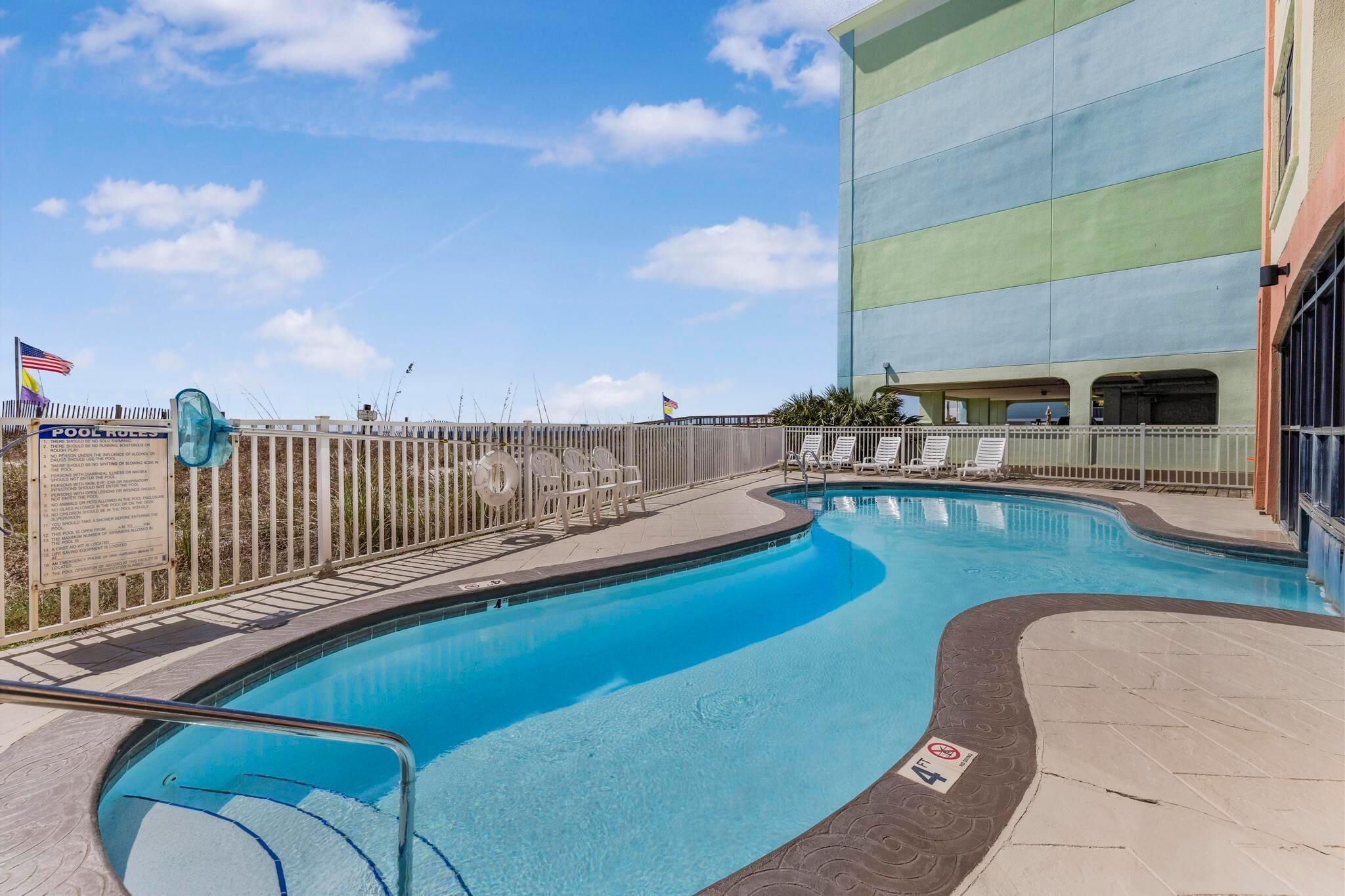 Une piscine entourée d’une clôture, avec des chaises longues et un panneau indiquant les règles d’utilisation de la piscine.