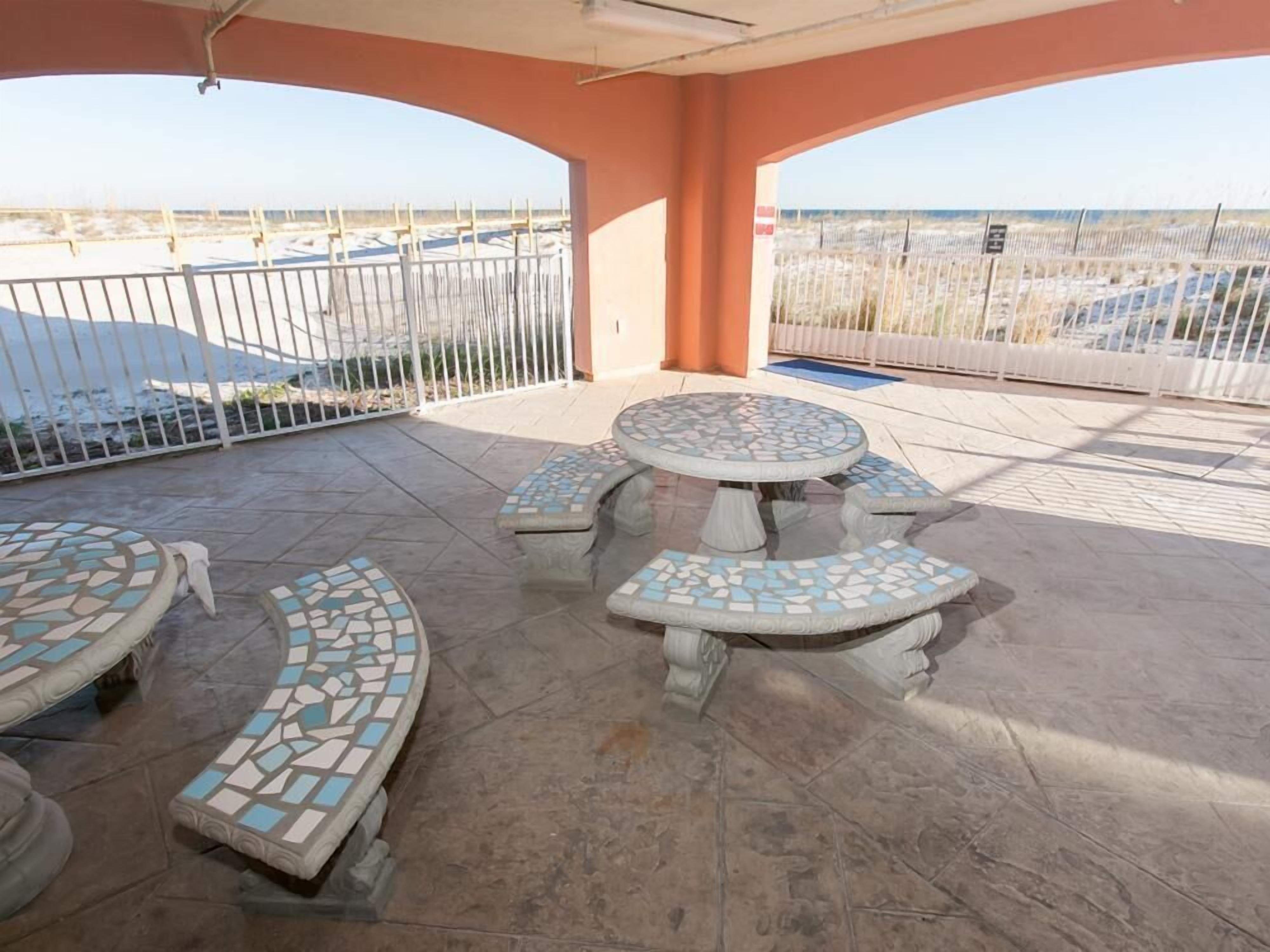 Un patio al aire libre con mesas y bancos de mosaico, una cerca blanca y vistas a una playa y coches aparcados.