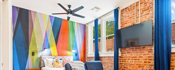 A room with a colorful geometric wall, a ceiling fan, a TV mounted on a brick wall, and a window with blue curtains.