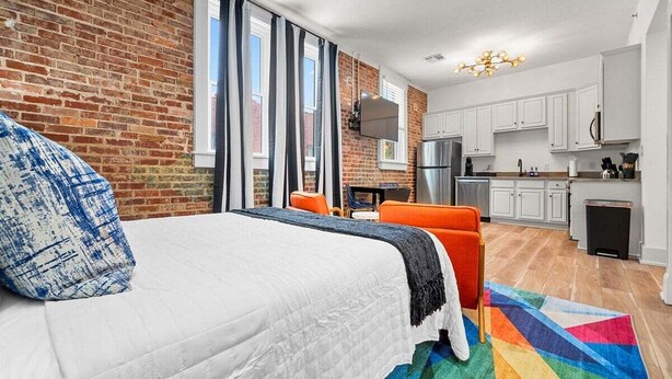 A modern hotel room with a brick wall, a bed with a white duvet, a colorful rug, a small kitchen area with white cabinets, and a TV mounted on the wall. - Funky Bungalow Studio · Walk to Bathhouse Row (Hot Springs)