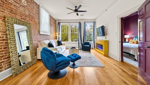 A room with a brick wall, a ceiling fan, a sofa, a blue chair, a small table, and a bed in the background. - Vibrant Loft Steps from Bathhouse Row (Hot Springs)