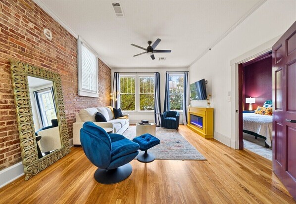 A room with a brick wall, a ceiling fan, a sofa, a blue chair, a small table, and a bed in the background.