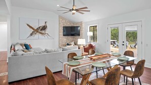 A modern living room with a sofa, dining table, chairs, a television, and a stone accent wall. - 6 BR Private Ranch, 2 Hot Tubs, Game Barn (Fredericksburg)