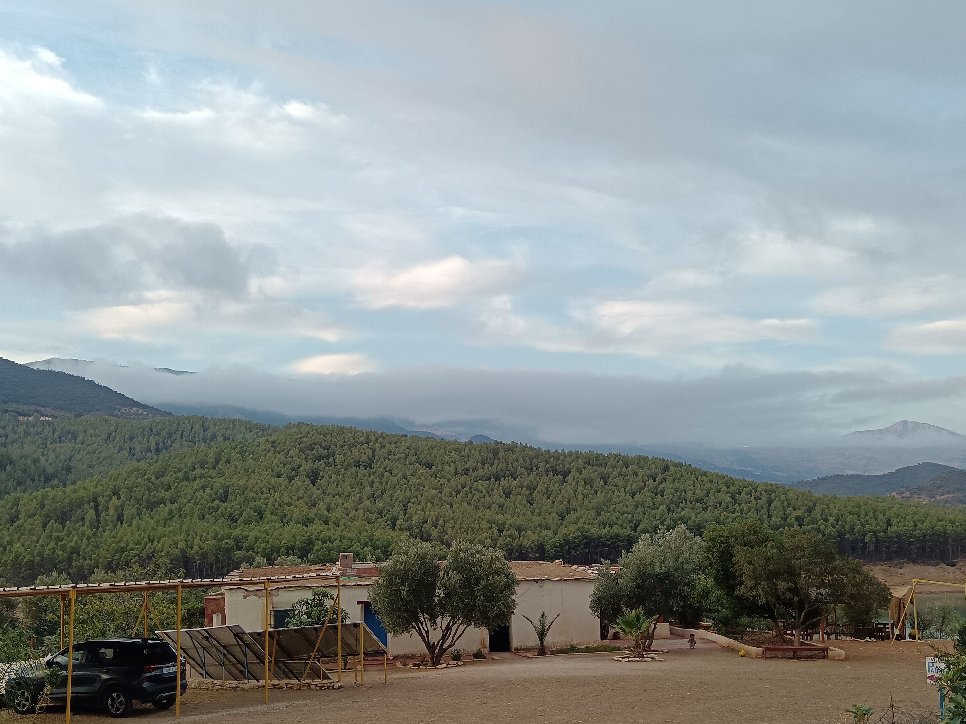 A rural landscape with a house, a parked car, and a clear view of the surrounding forest and hills.