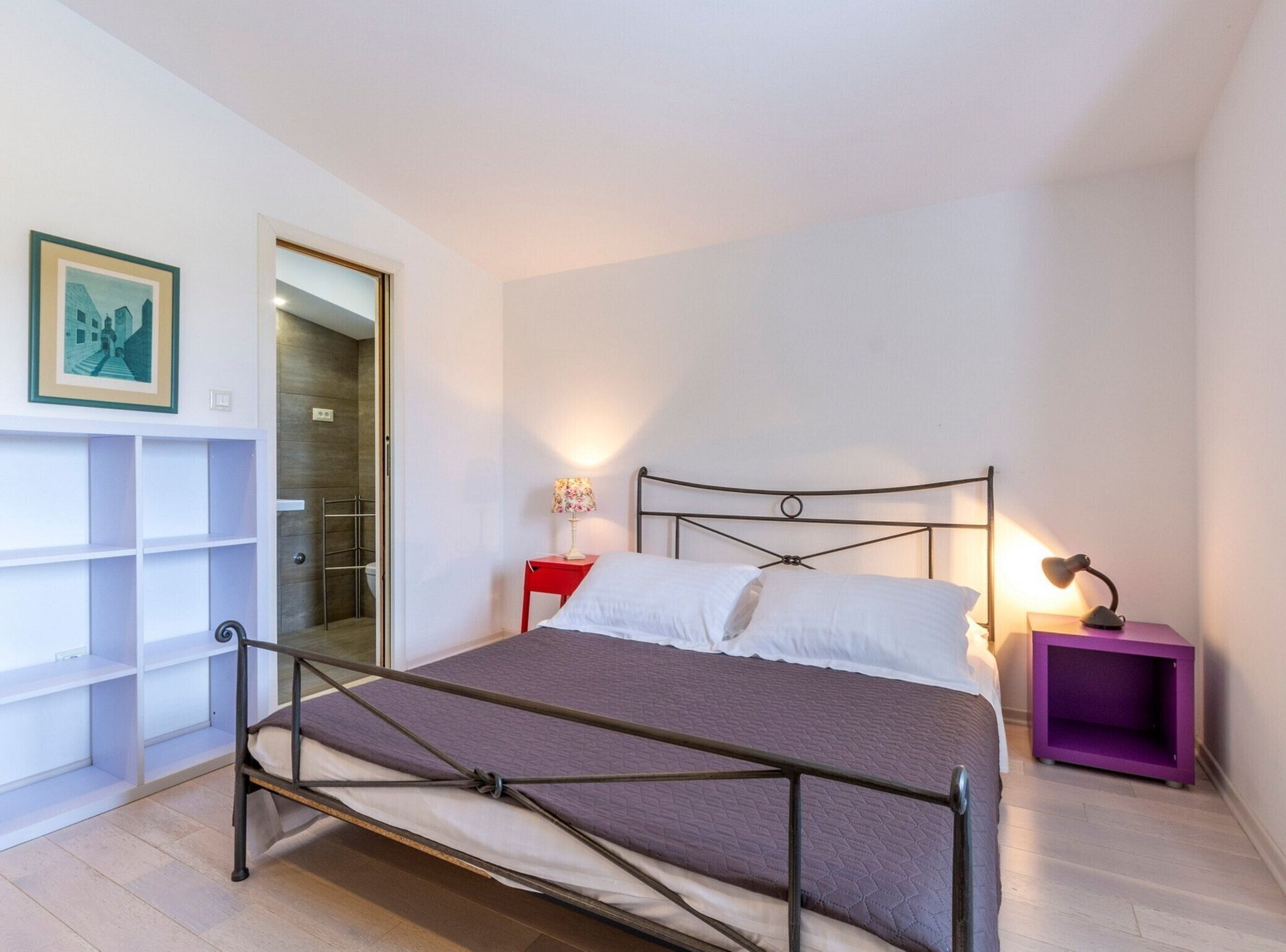 Een slaapkamer met een metalen bedframe, een rode nachtkastje, een paars bijzettafeltje en een lijst met een afbeelding aan de muur.