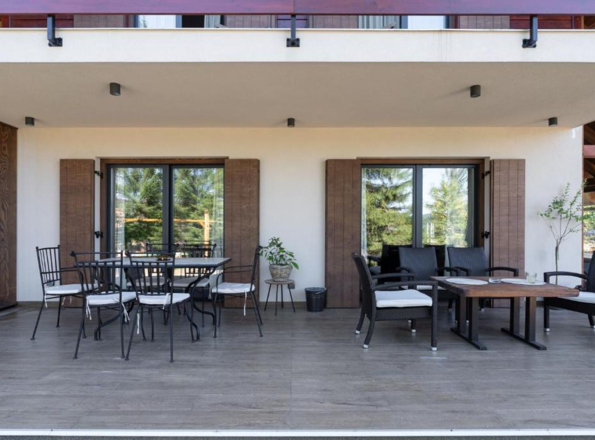 An outdoor patio with a dining table and chairs, large windows, and a view of greenery.