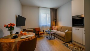 Un saló modern amb un sofà, una taula de dinar per a dues persones, un televisor penjat a la paret i una zona de cuina amb forn.