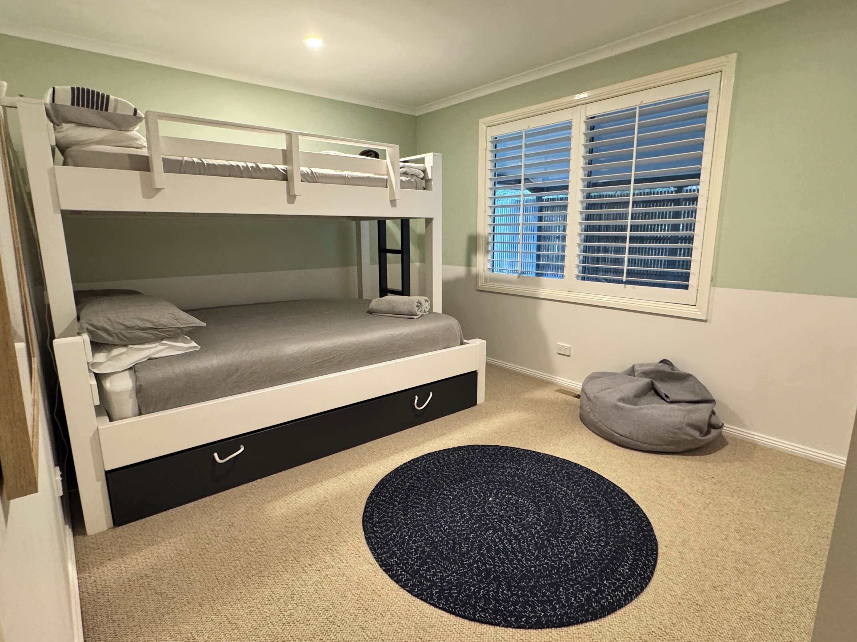 A bunk bed with a desk drawer, a window with blinds, a grey bean bag, and a circular rug.