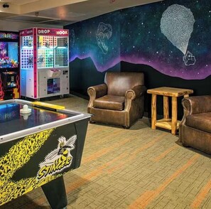 Une table de billard ornée du logo « Stinger », des jeux d’adresse à pinces, un fauteuil en cuir marron et une petite table en bois, le tout dans une pièce dont le mur est décoré d’une fresque sur le thème de l’espace.