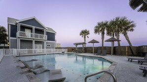 A swimming pool with lounge chairs and umbrellas, a wooden fence, and a building with balconies. - Walk to Beach~Heated Pool~BBQ Area~Balcony~Island (Corpus Christi)