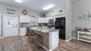 A modern kitchen with white cabinetry, a granite countertop island, and nautical-themed wall decor. - Walk to Beach~Heated Pool~BBQ Area~Balcony~Island (Corpus Christi)