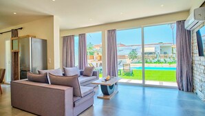 A spacious living room with a grey sofa, a coffee table, and a large sliding glass door leading to a pool area. - Villa Cinque Elementi by Estia (Ionian Islands)