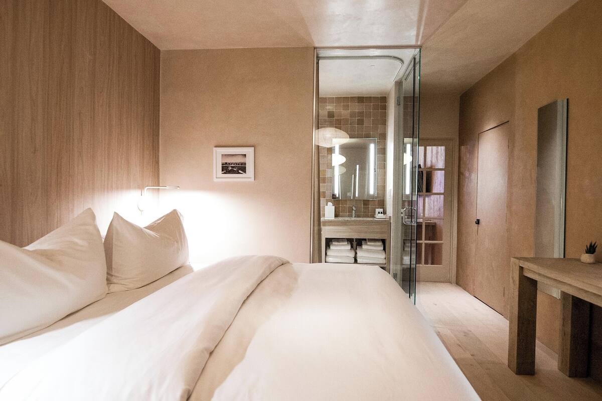 Une chambre à coucher moderne avec un grand lit, une table de chevet, un miroir et une salle de bain visible à travers une porte en verre.
