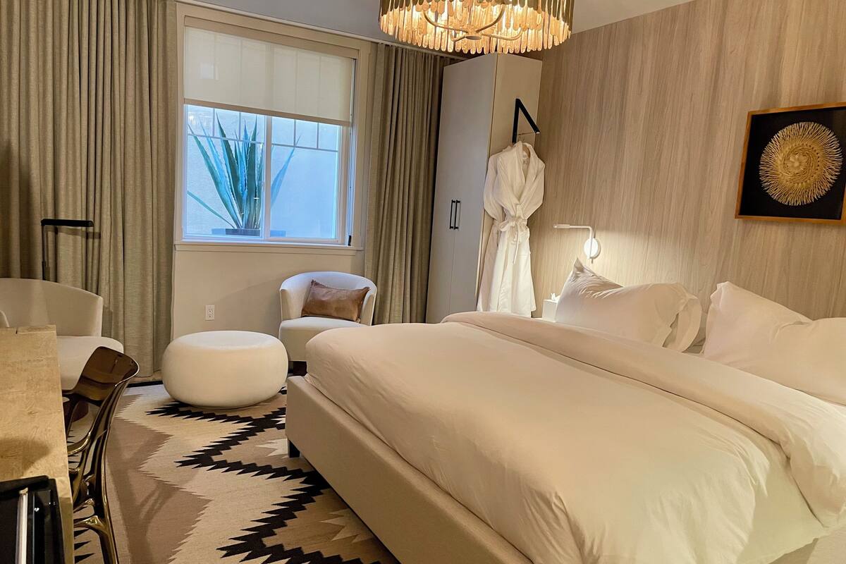 Une chambre d’hôtel moderne dotée d’un grand lit, d’un coin salon avec un fauteuil et un pouf, d’une fenêtre avec des rideaux et d’un élément décoratif mural.